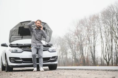 Sad driver calling car service, opening hood, having engine problem standing near broken car on the road. Car breakdown concept.