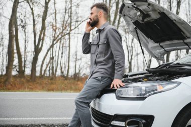 Sad driver calling car service, opening hood, having engine problem standing near broken car on the road. Car breakdown concept.