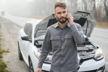 Sad driver calling car service, opening hood, having engine problem standing near broken car on the road. Car breakdown concept.
