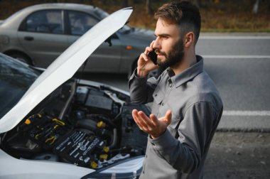 Sad driver calling car service, opening hood, having engine problem standing near broken car on the road. Car breakdown concept.