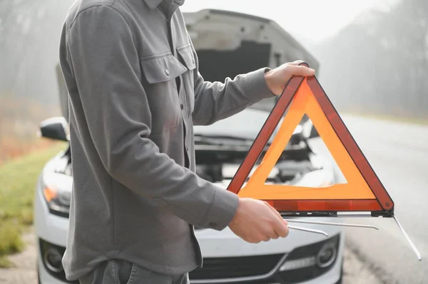 Young upset casual man trying to fix his broken car outdoors. Man waiting for towing service for help car accident on the road. Roadside assistance concept