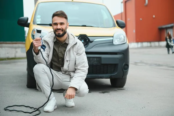 Man Holding Power Charging Cable For Electric Car In Outdoor Car Park. And he s going to connect the car to the charging station in the parking lot near the shopping center.