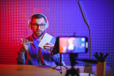 Genç erkek teknoloji blogcusu video blogu veya video blogu kaydediyor. Yeni akıllı telefon ve diğer cihazlar hakkında. Blog yazma, ev işleri konsepti. Masadaki aletlere odaklan. Web Afişi.