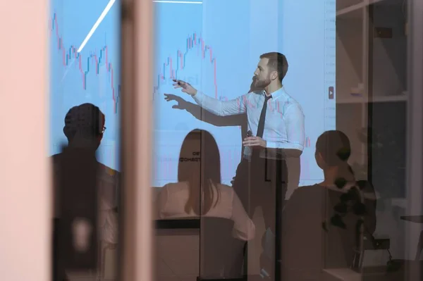 Young Businessman Explaining Graphs To His Colleagues On Projector.