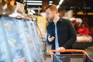 Süpermarkette: yakışıklı adam akıllı telefon kullanan ve beslenme bakar konserveler değerini. O ile alışveriş sepeti konserve ürünleri bölümünde duruyor