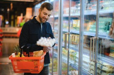 Süpermarketteki müşteri. Market alışverişi yapan bir adam, araba ile ayakta duruyor, içeride yiyecek seçiyor. Gıda mağazasından alışveriş yapan adam. Seçici Odaklanma, Boşluğu Kopyala