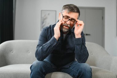 Üzgün, sorunlu orta yaşlı bir adam kanepede oturuyor, arkadaşlarını özlüyor, kötü hissediyor sağlıksız, evde depresyondan muzdarip..