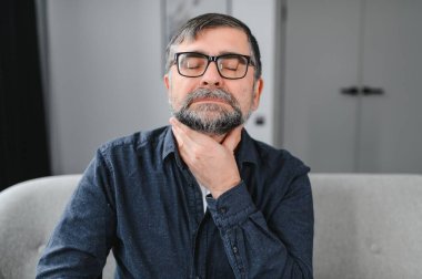 Kıdemli adam soğuktan muzdarip. Boğazı ağrıyan yaşlı bir adamın fotoğrafı. Yaşlı beyaz adam boğazına dokunuyor. Boğaz ağrısı, bademcik iltihabı ya da tiroit bezi sorunu. Yaşlı adam kendini iyi hissetmiyor.