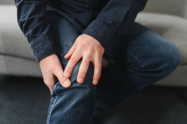 Yaşlı adam evdeki koltukta oturuyor, diz eklemleri ağrıyor, bacağını tutuyor, osteoartrit konsepti