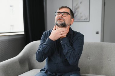 Kıdemli adam soğuktan muzdarip. Boğazı ağrıyan yaşlı bir adamın fotoğrafı. Yaşlı beyaz adam boğazına dokunuyor. Boğaz ağrısı, bademcik iltihabı ya da tiroit bezi sorunu. Yaşlı adam kendini iyi hissetmiyor.