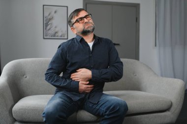 Mutsuz yaşlı adam mide ağrısı, dayanılmaz acıdan karnına bastıran adam, midesi ağrıyan olgun adam oturma odasında kendini rahatsız hisseden adam..