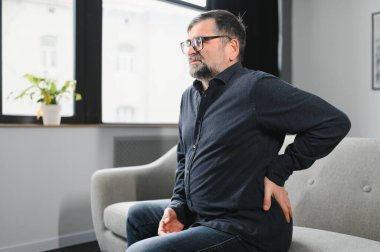 İnsanlar, Sağlık ve Tedavi Bölümü. Akut sırt ağrısından muzdarip hasta olgun bir adamın portresi, evdeki koltukta oturan, sırtının alt kısmına elle dokunan biri. Sağlık Sorunu Konsepti.