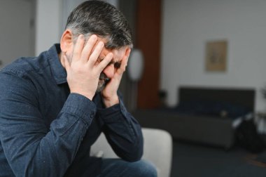 Orta yaşlı, kır saçlı, stresli bir adam evdeki koltukta oturuyor..