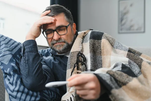 Sick senior man using thermometer checking his temperature suffering from seasonal flu or cold