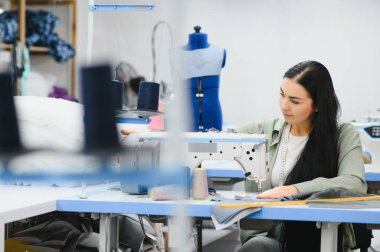 Cheerful young seamstress sits at the table with working things, in atelier.