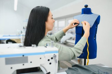 Cheerful young seamstress sits at the table with working things, in atelier.