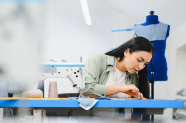 Cheerful young seamstress sits at the table with working things, in atelier.