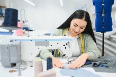 Cheerful young seamstress sits at the table with working things, in atelier.