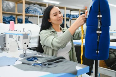 Cheerful young seamstress sits at the table with working things, in atelier.