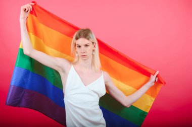 Young gay man 20s with make up wrapped in rainbow striped flag isolated on background, studio portrait. People lifestyle fashion lgbtq concept