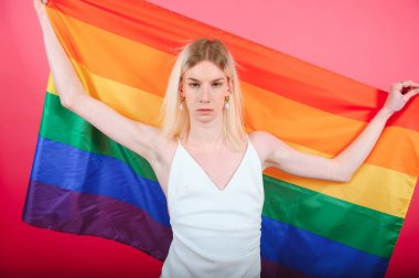 Young gay man 20s with make up wrapped in rainbow striped flag isolated on background, studio portrait. People lifestyle fashion lgbtq concept
