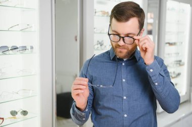 Satisfied Customer. View of happy young male client wearing new glasses, standing near rack and showcase with eyewear. Smiling man trying on spectacles.