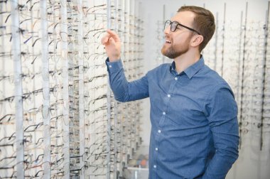 Satisfied Customer. View of happy young male client wearing new glasses, standing near rack and showcase with eyewear. Smiling man trying on spectacles.