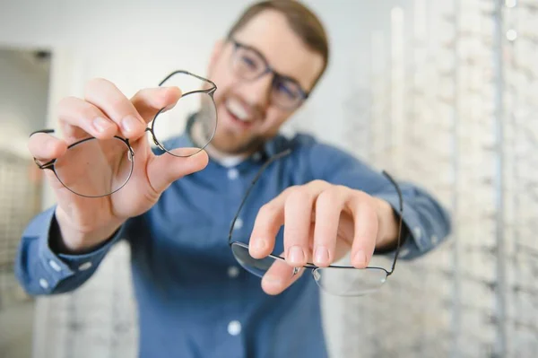 In Optics Shop. Portrait of male client holding and wearing different spectacles, choosing and trying on new glasses at optical store. Man picking frame for vision correction, closeup