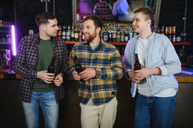 Young men in casual clothes are talking, laughing and drinking while sitting at bar counter in pub.