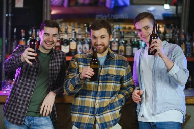 Young men in casual clothes are talking, laughing and drinking while sitting at bar counter in pub.