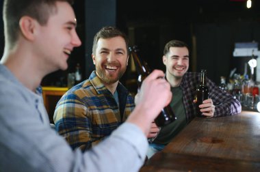 Three young men in casual clothes are smiling, holding bottles of beer while standing near bar counter in pub.