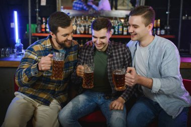 Young men in casual clothes are talking, laughing and drinking while sitting at bar counter in pub.