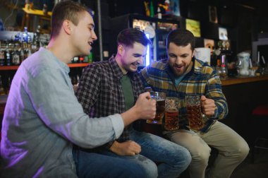 Young men in casual clothes are talking, laughing and drinking while sitting at bar counter in pub.