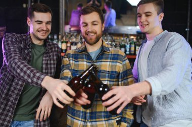 Three young men in casual clothes are smiling, holding bottles of beer while standing near bar counter in pub.