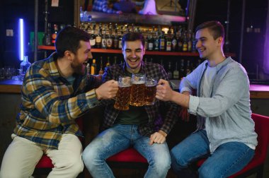 Young men in casual clothes are talking, laughing and drinking while sitting at bar counter in pub.