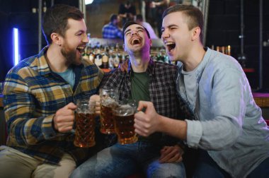 Young men in casual clothes are talking, laughing and drinking while sitting at bar counter in pub.
