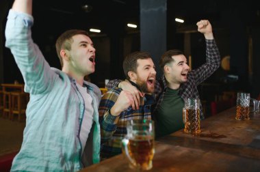 Soccer fans cheering for favourite team while enjoying drinks. Group of supporters celebrating victory while watching football match on tv in a club. Happy friends shouting while cheering for a goal