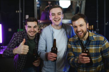 Three young men in casual clothes are smiling, holding bottles of beer while standing near bar counter in pub.
