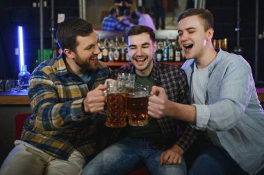 Young men in casual clothes are talking, laughing and drinking while sitting at bar counter in pub.