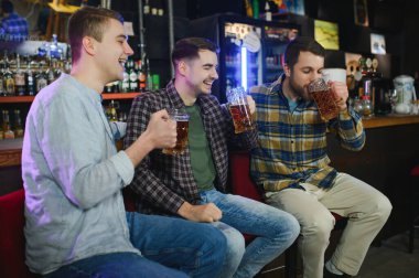 Young men in casual clothes are talking, laughing and drinking while sitting at bar counter in pub.