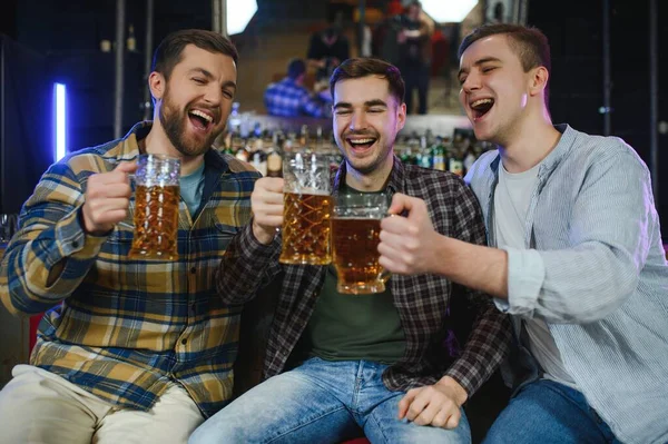 Old friends meeting. Happy young men in casual wear toasting with beer while sitting in beer pub together.