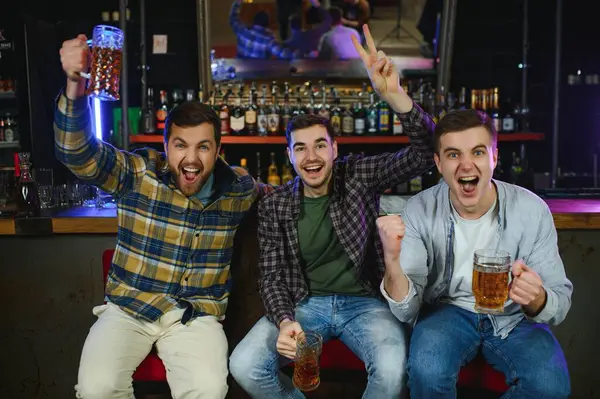 Soccer fans cheering for favourite team while enjoying drinks. Group of supporters celebrating victory while watching football match on tv in a club. Happy friends shouting while cheering for a goal