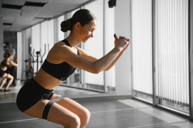 Motivation for girls. Modern young woman in sport clothing crouching using resistance band while exercising in the gym