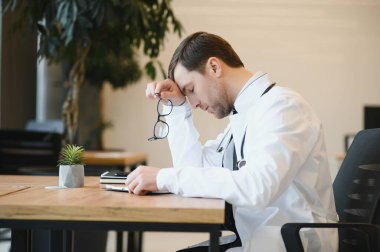 Stressed male doctor sat at his desk. Mid adult male doctor working long hours. Overworked doctor in his office. Not even doctors are exempt from burnout