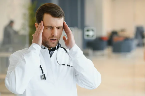 Handsome young doctor in white coat is touching his temples while standing in hospital hall