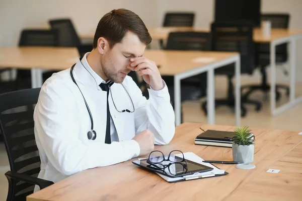 Stressed male doctor sat at his desk. Mid adult male doctor working long hours. Overworked doctor in his office. Not even doctors are exempt from burnout