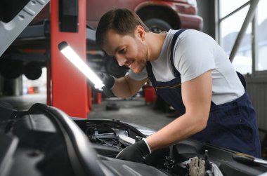 A mechanic is doing car checkup under the hood with flashlight at mechanic's shop