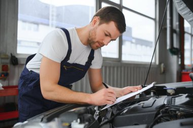 car mechanic writing while holding clipboard near cars.