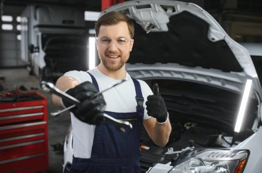 With tool in hand. Adult man in colored uniform works in the automobile salon.