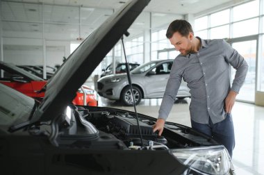 Looks under the hood of automobile. Young man in the car dealership